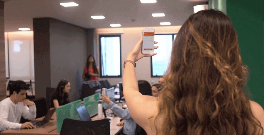 pessoa vista de costas em escritório movimentado suspendendo celular com a mão simulando bater ponto de forma móvel