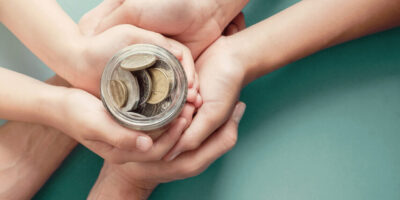 Pote de moedas sobre as mãos sobrepostas de dois adultos e uma criança