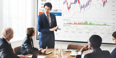 profissional em provável cargo de CFO apresentando dados em pé em frente a colegas sentados à mesa