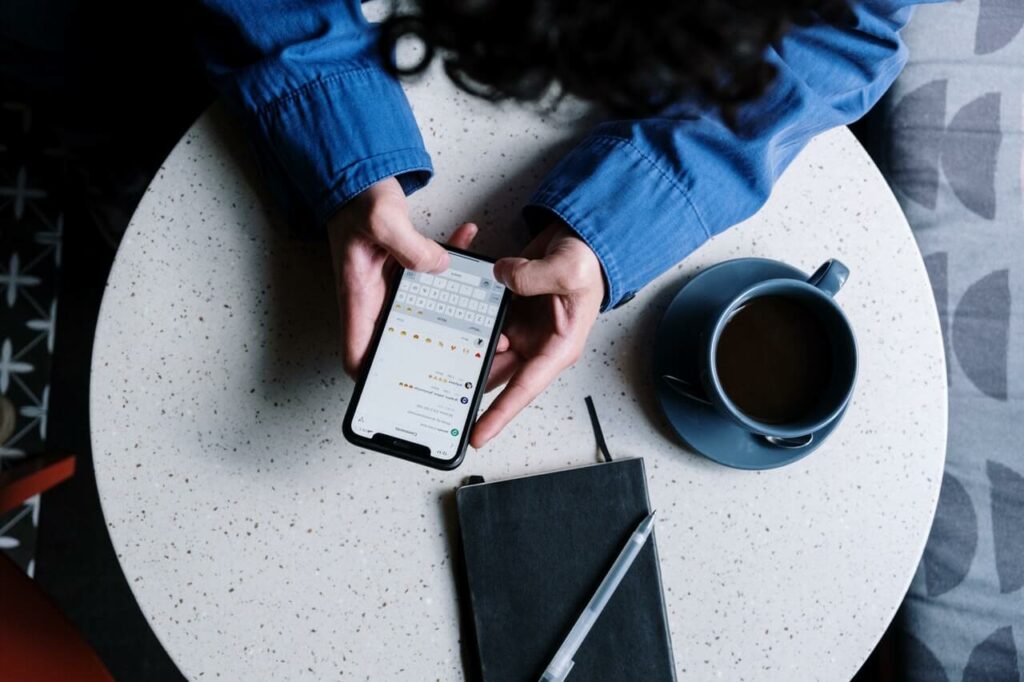 Mãos de uma pessoa segurando celular com rede social aberta sobre uma mesa em que há xícara, agenda e caneta.