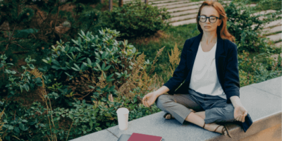 pessoa de roupa social com olhos fechados, pernas cruzadas e mãos sobre joelhos sentada em frente a notebook fechado simulando demissão silenciosa