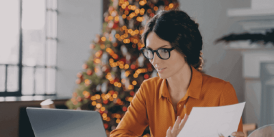 mulher de roupa social é óculos de grau sentada em frente a notebook aberto com folha de papel na mão e árvore de natal ao fundo simulando fim de ano no rh.