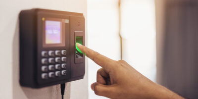 Imagem de um relógio de ponto biométrico posicionado numa parede. O dedo indicador de uma pessoa está posicionado no local onde a biometria digital é colhida.