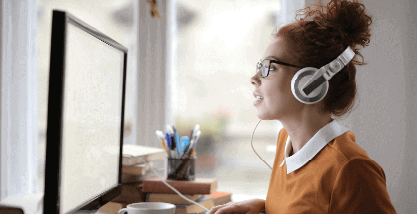 imagem de mulher sentada em frente a computador de mesa olhando para a tela e com fones de ouvido representando um possível acesso a um portal do colaborador.