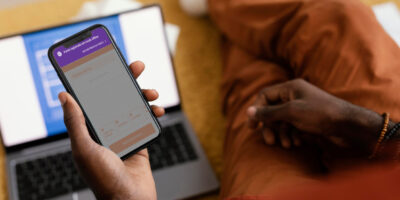 Imagem de celular com tela mostrando registro de ponto offline sendo segurado pela mão esquerda de uma pessoa. Ao fundo se vê o corpo da pessoa, que estã sentada ao lado de notebook.