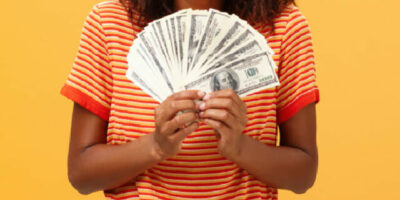 imagem de mulher sorrindo de boca aberta enquanto segura várias notas de dinheiro em leque nas duas mãos, na altura do peito.