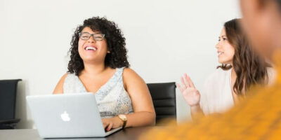 Mulher ri em frente a um notebook enquanto conversa com outra, numa ideia de qualidade de vida da jornada de trabalho não linear